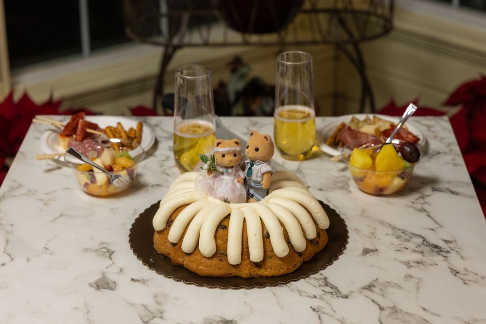 Bundt cake with white frosting on marble table, flanked by wine glasses and fruit bowls with decorative figurines.