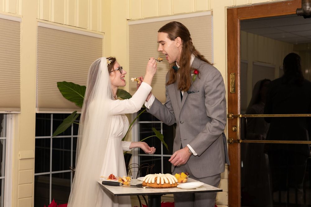 Bride and groom sharing wedding cake at reception table, bride in white veil feeding groom in gray suit.