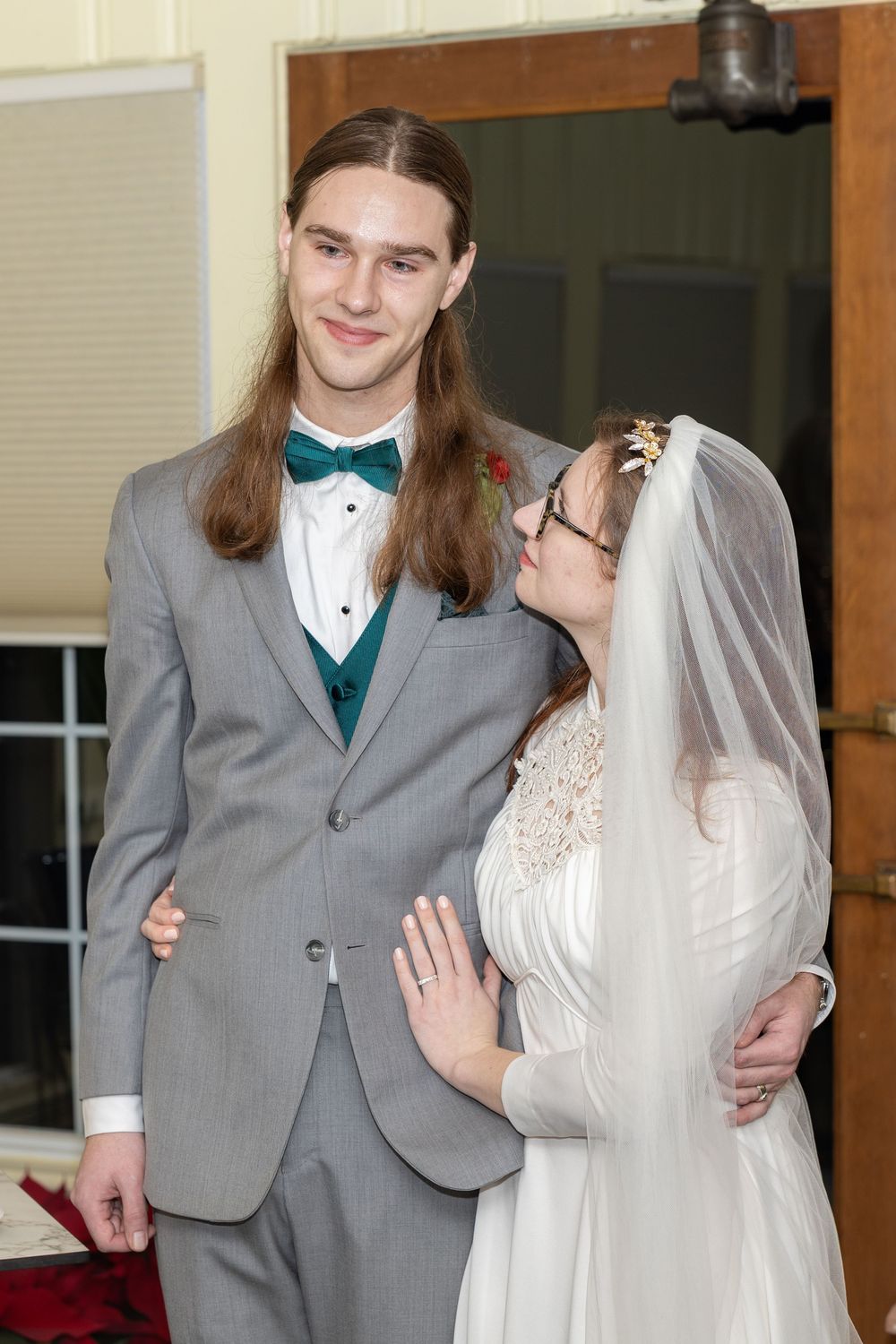 A bride in white dress and veil stands beside groom in gray tuxedo with teal bow tie at wedding ceremony.