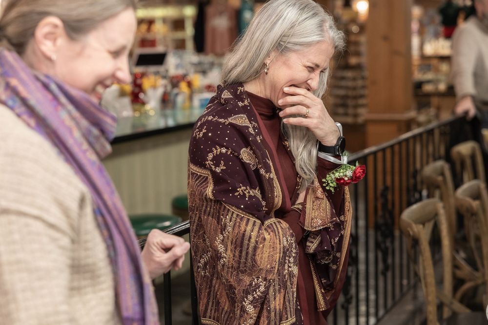 Two women in a rustic indoor setting, one with gray hair wearing a floral shawl, laughing warmly.
