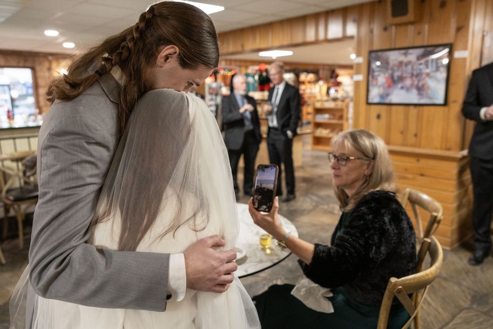 Bride in white gown and veil gets assistance from two women in a cozy wood-paneled shop setting.