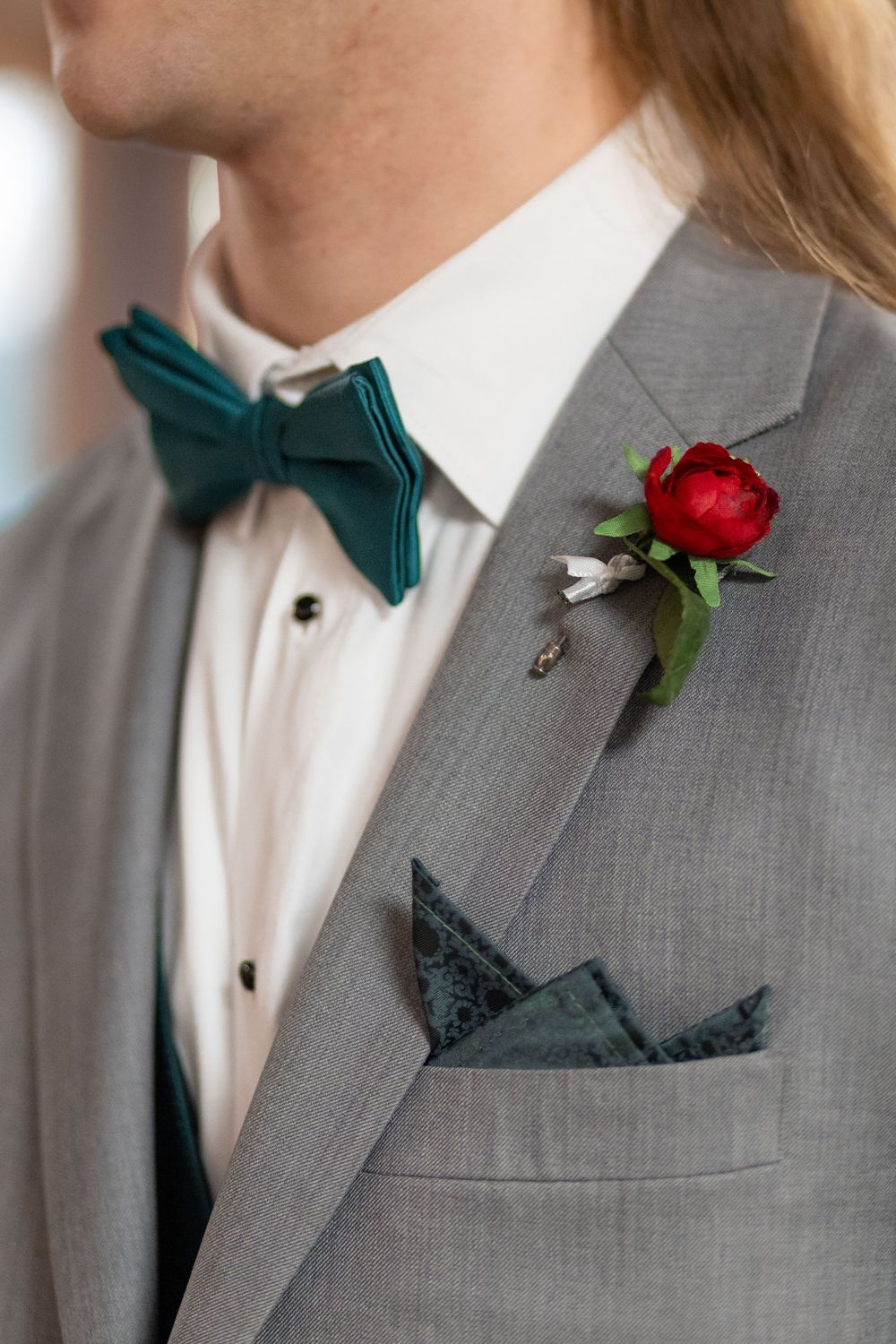 Groom in gray suit with teal bow tie, red peony boutonniere, and teal pocket square at wedding.
