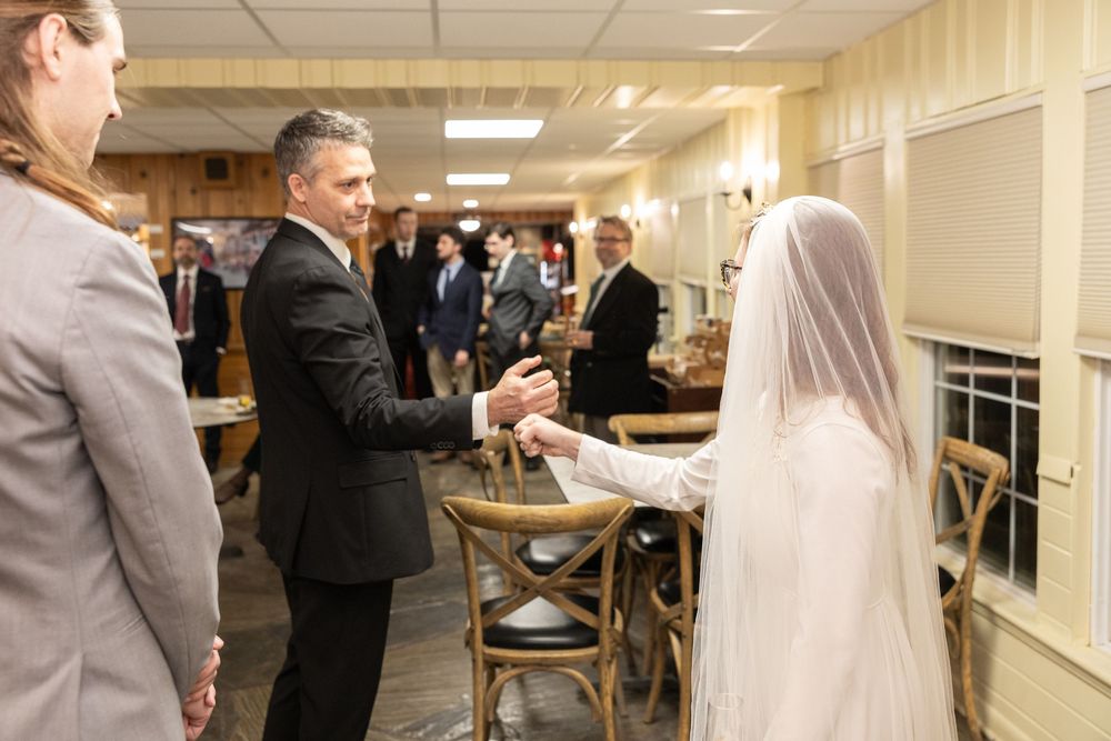 A groom in a black suit holds hands with a bride in a white veil during a wedding reception indoors.