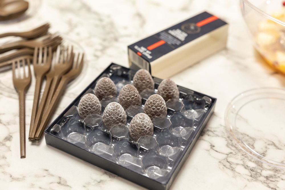 Chocolate pinecone truffles displayed in a black box on marble surface with gold cutlery and packaging nearby.