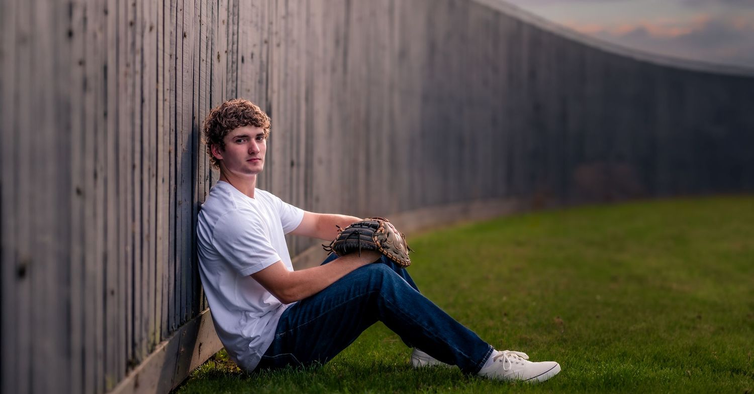 Teen boy in letterman jacket holds baseball glove on field near scoreboard during senior portrait session.
