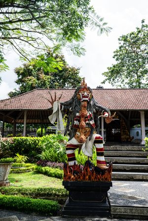 Colorful Balinese Hindu deity statue with traditional ornaments stands in a lush temple garden with traditional architecture.