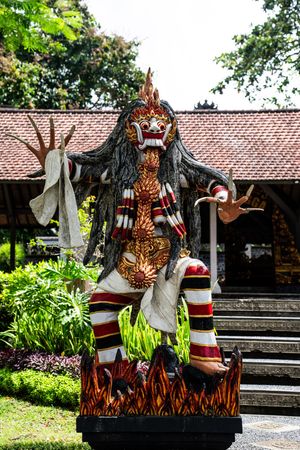 Traditional Balinese demon statue with colorful costume and fierce expression displayed outdoors near a temple garden.