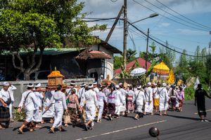 Balinese Hindu procession with people in white traditional attire carrying ceremonial offerings along a sunny street.