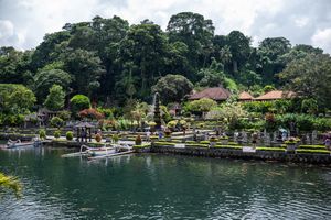 Lush tropical garden with ornamental pools, traditional Balinese architecture, and dense green forest backdrop on a cloudy day.