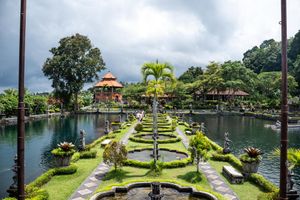 Lush Balinese water palace garden with manicured hedges, koi ponds, and traditional pavilion under dramatic cloudy skies.