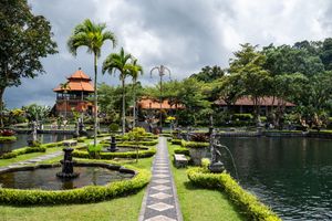 Lush Balinese water palace garden with palm trees, manicured hedges, fountains, and traditional architecture under cloudy skies.