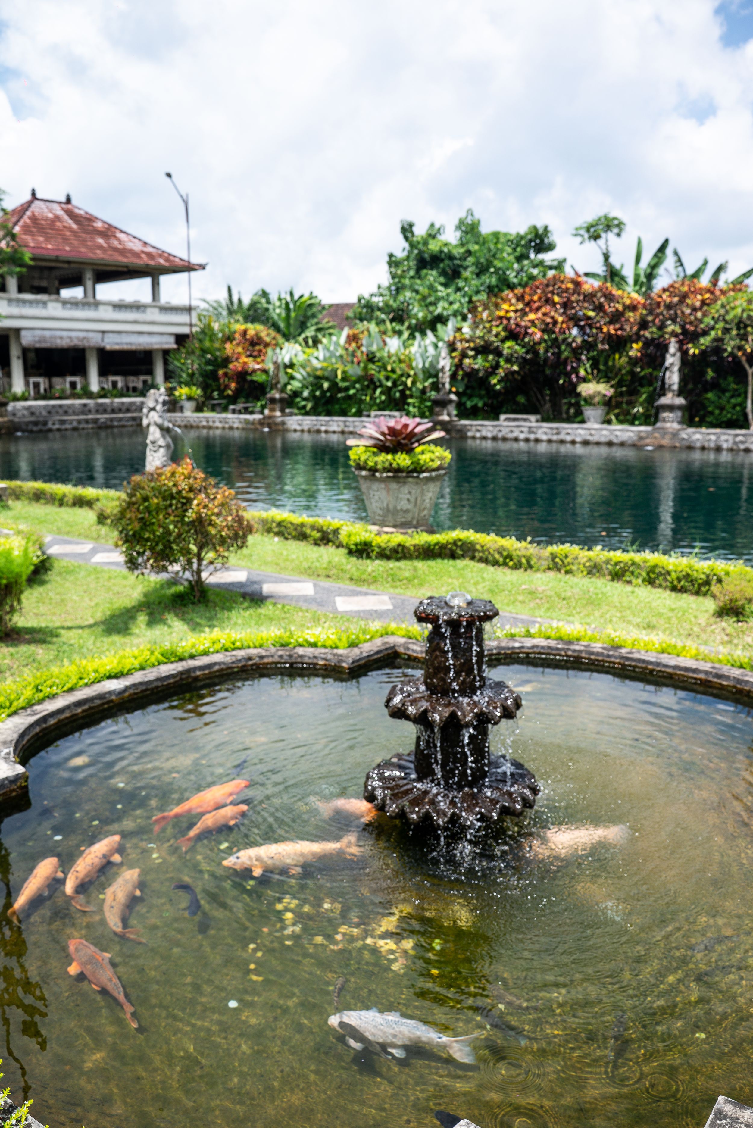 Koi fish swim in a ornate stone fountain at a lush Balinese garden with a traditional pavilion.