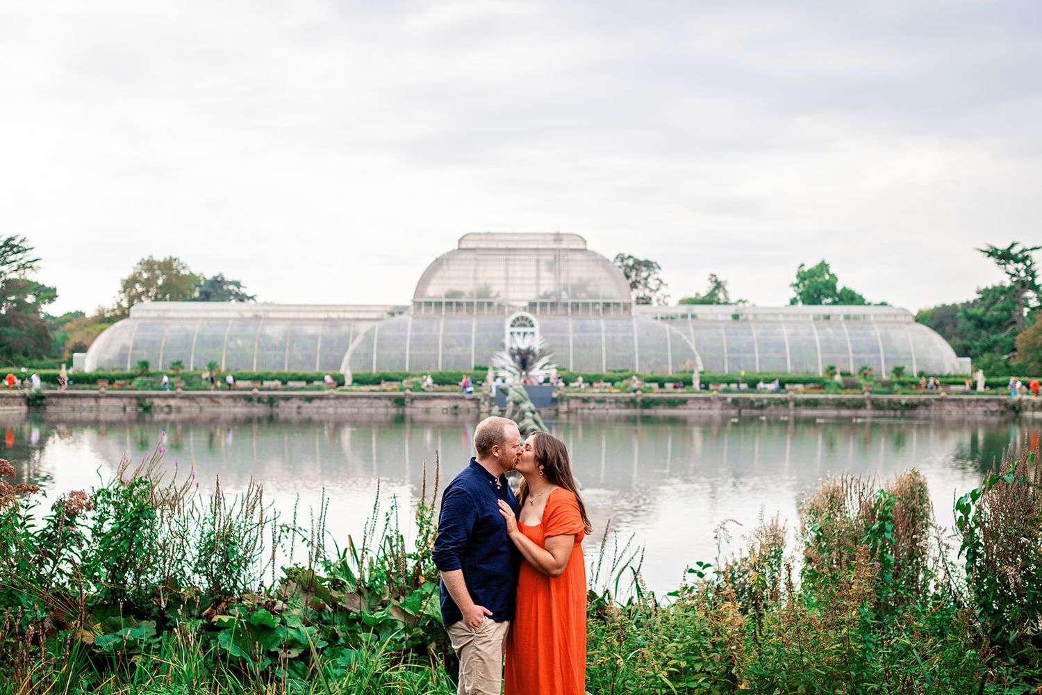 Couple kissing in front of Kew Gardens Palm House greenhouse and lake, woman in orange dress, man in navy shirt.