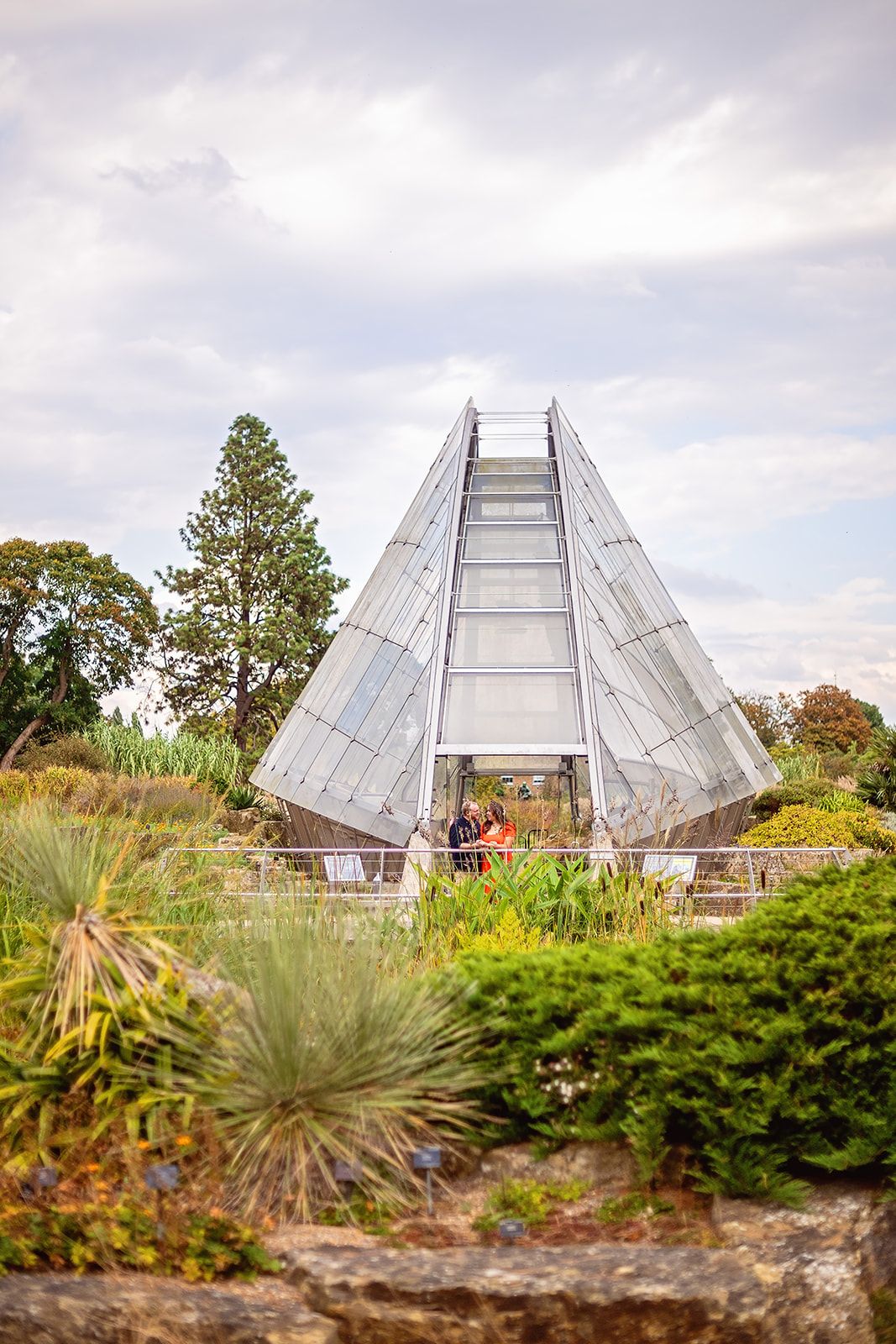 A geometric glass pyramid greenhouse structure surrounded by lush garden plants under a cloudy sky.