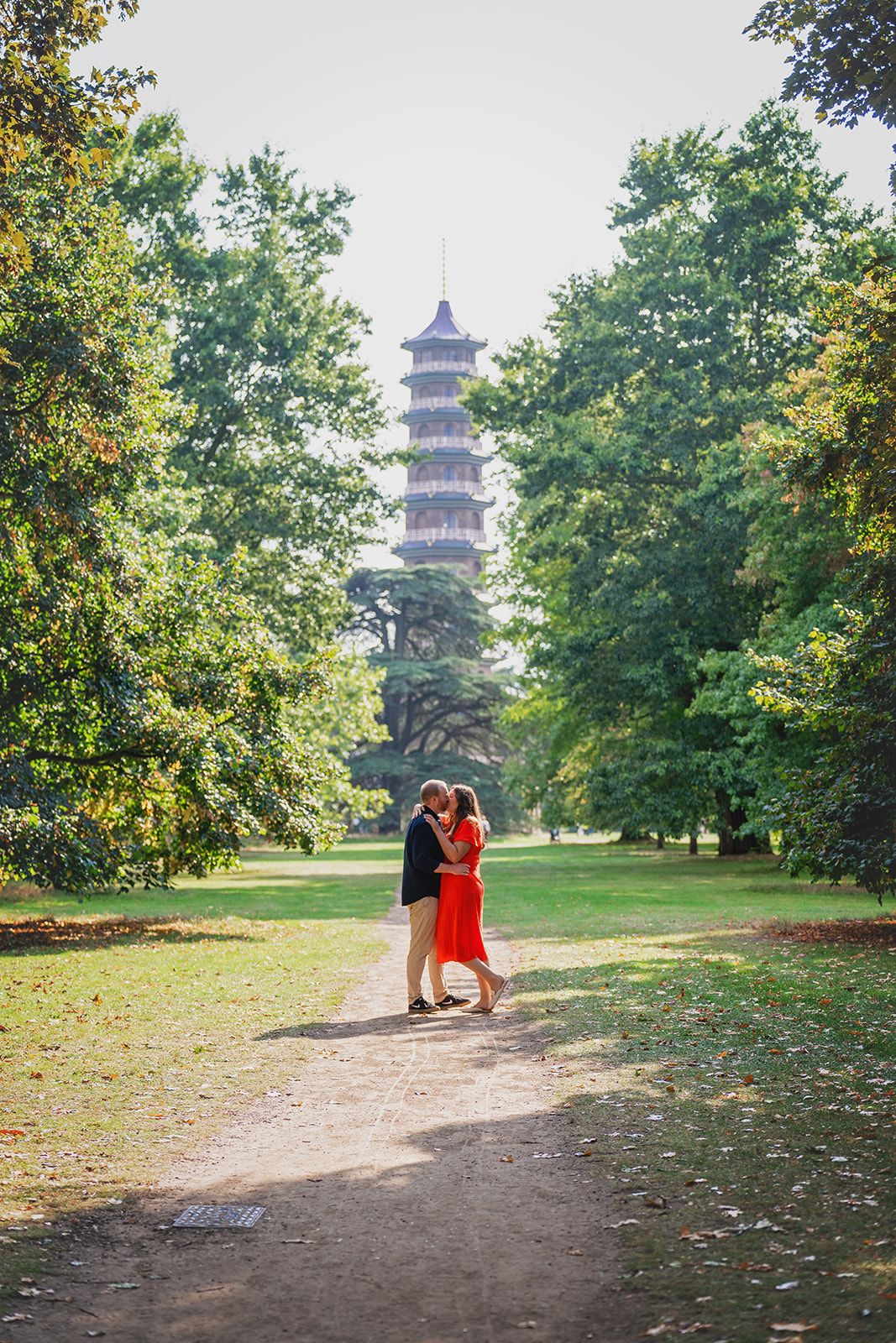 Couple embracing on a park path with a pagoda tower visible through lush green trees in the background.