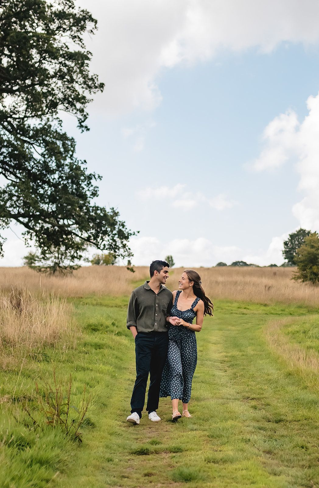 A couple walks hand-in-hand through a lush green meadow path, smiling at each other beneath a partly cloudy sky.