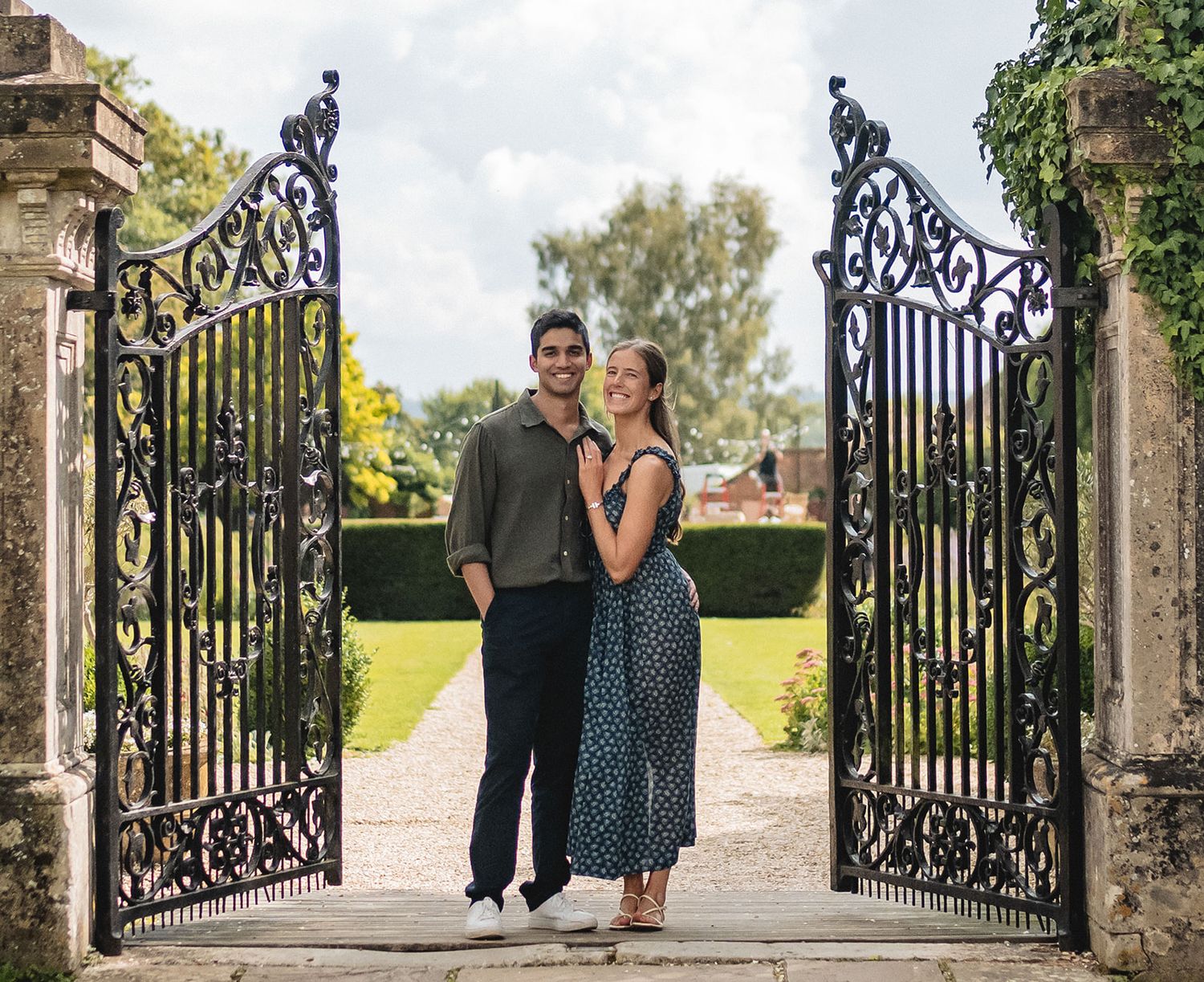 Couple smiling and embracing in ornate black iron garden gates with lush green grounds behind them.