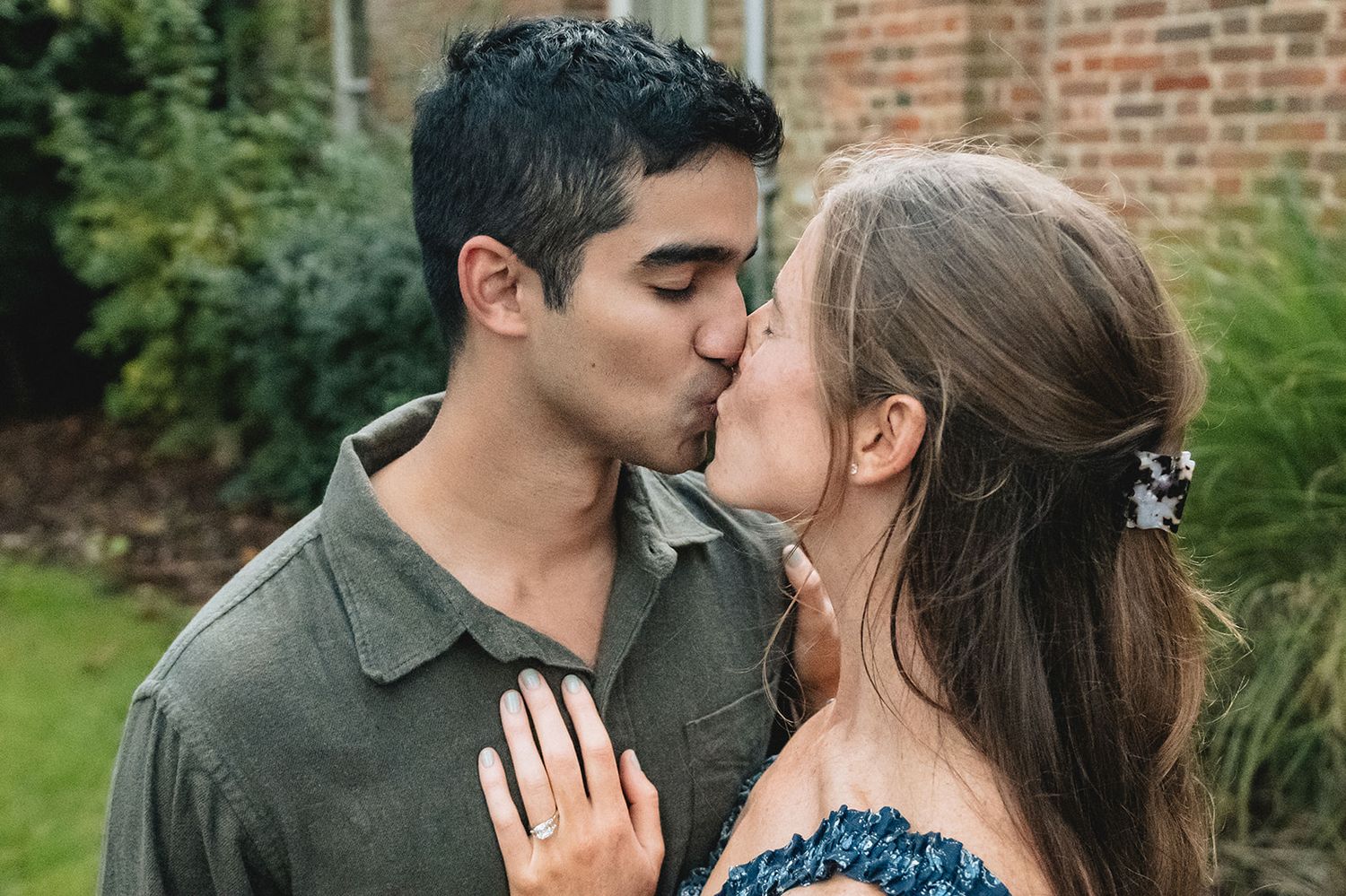 A couple shares a romantic kiss outdoors, with the woman wearing a floral accessory in her hair.