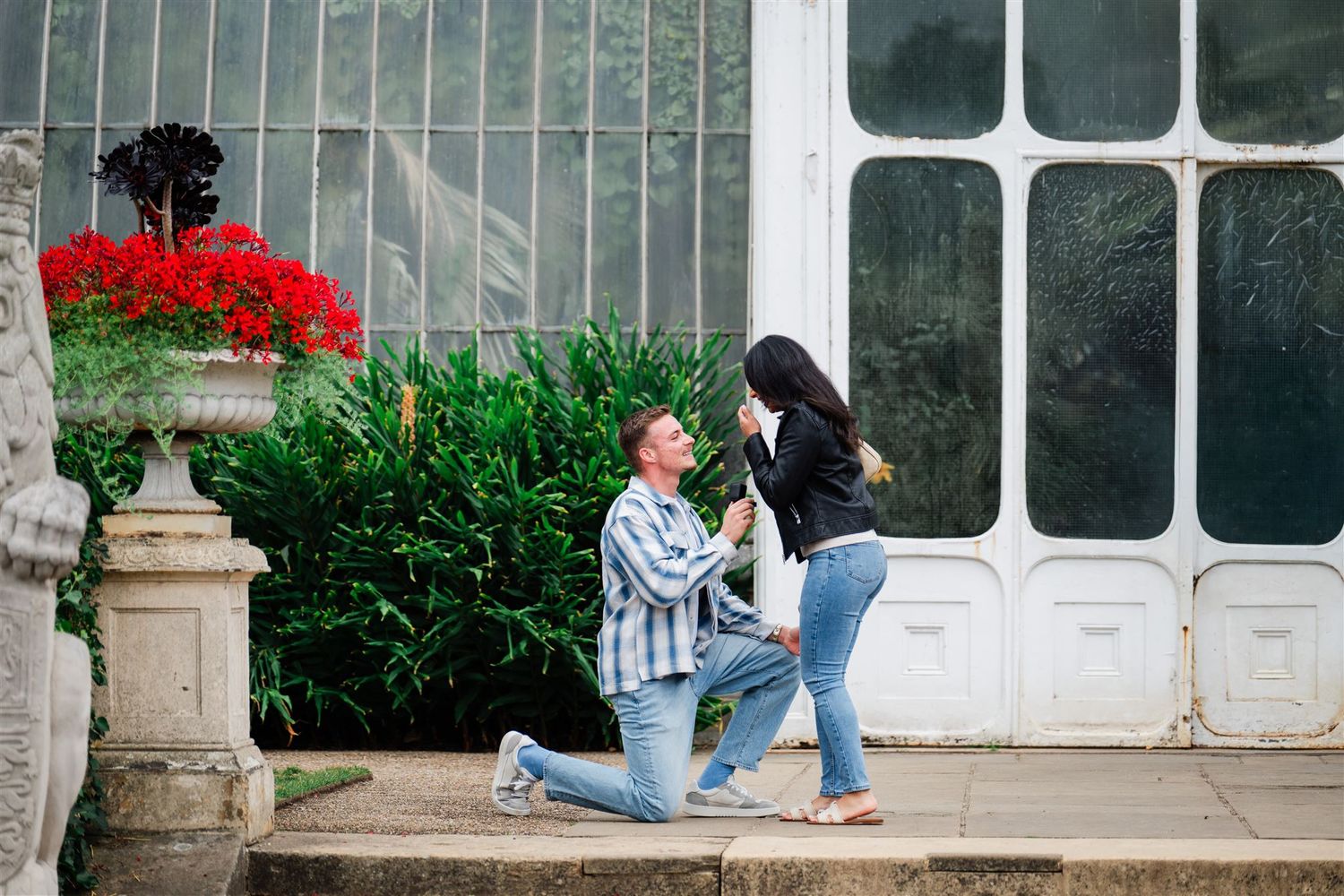 Man proposing on one knee to woman outdoors near red flowers and greenhouse.