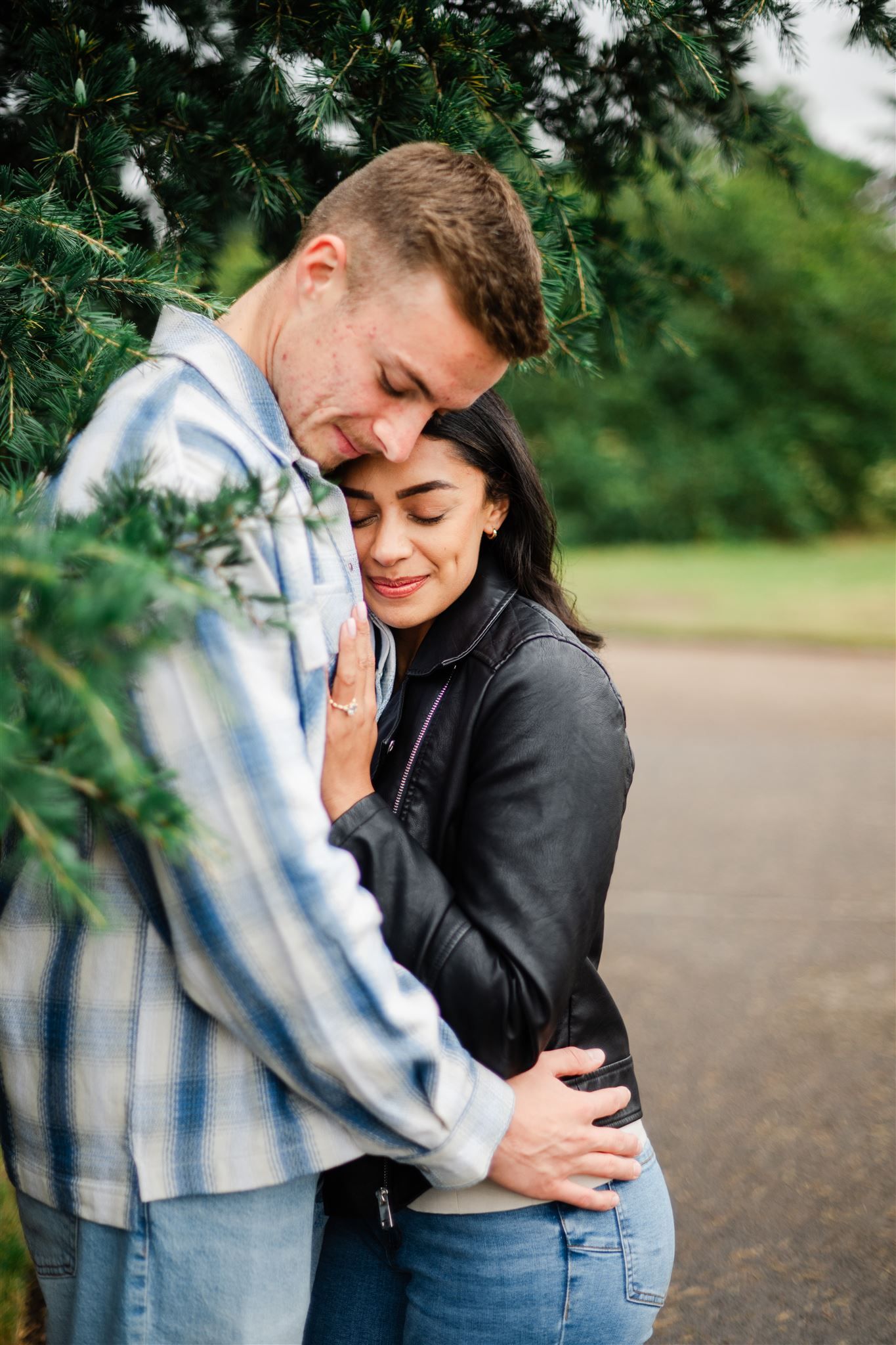 A couple embraces tenderly under evergreen trees, the woman in a leather jacket leaning into a man in a plaid shirt.