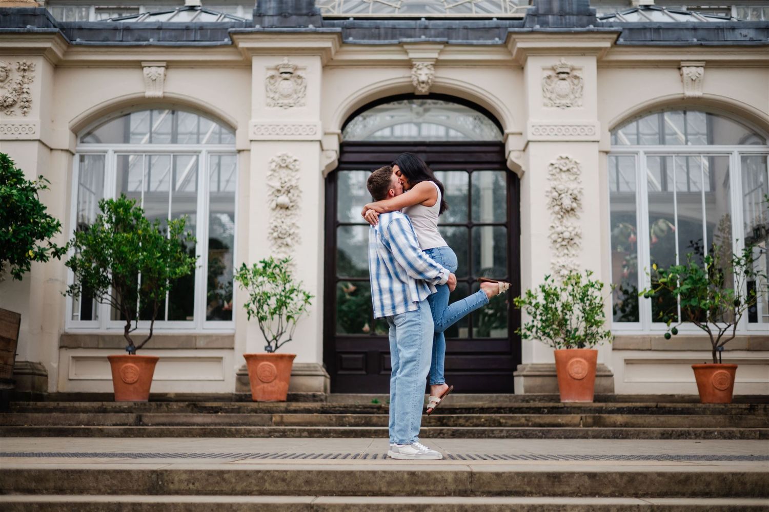 A couple embraces joyfully on steps before an elegant glass conservatory building.