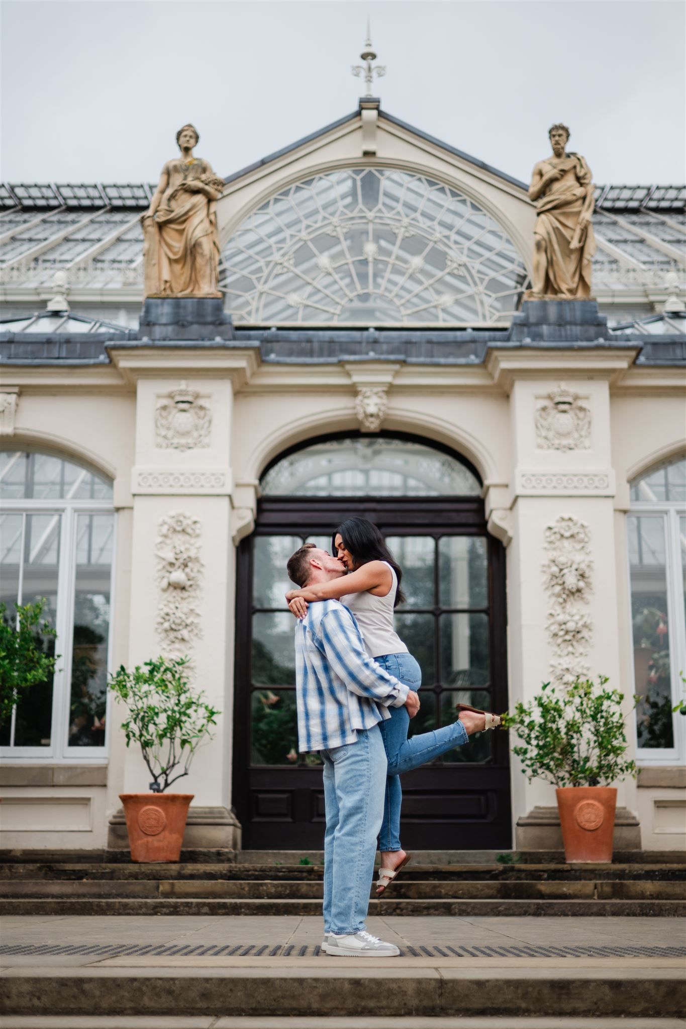 A couple shares a romantic lift and kiss in front of an ornate classical conservatory.