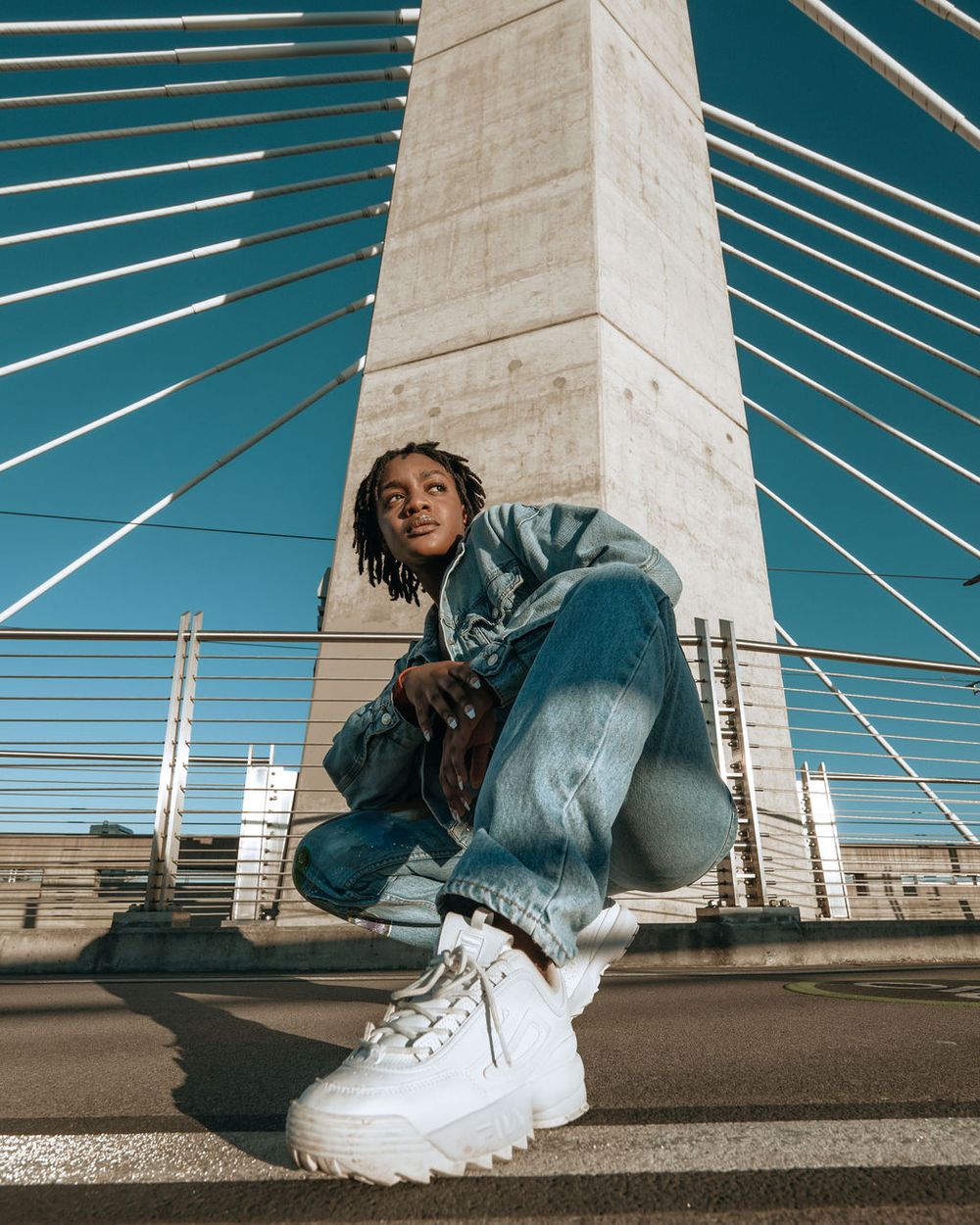 Wide angle shot from below showing someone crouching on a cable bridge wearing baggy jeans and white platform sneakers in Portland, OR.