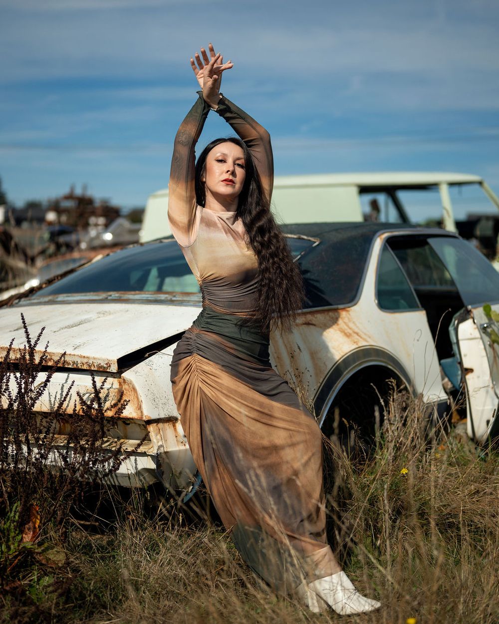 A dance performer in a flowing brown dress poses gracefully against a car at sunset in an urban setting in chic editorial photography.