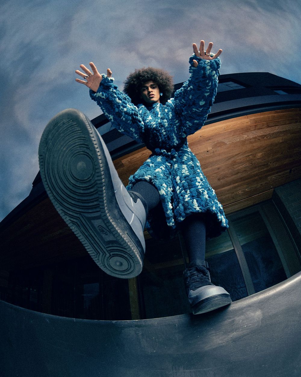 Person in blue floral coat with afro hair raises hands dramatically, large sneaker in foreground, fisheye lens perspective.
