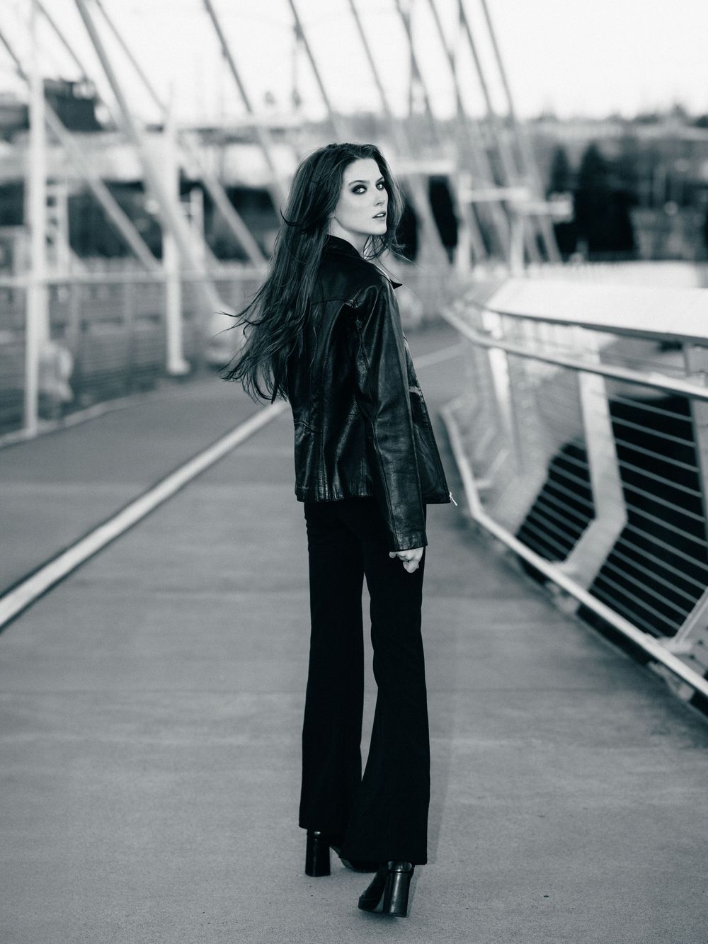 Black and white photo of a young woman in a leather jacket and flare pants standing on a modern bridge.
