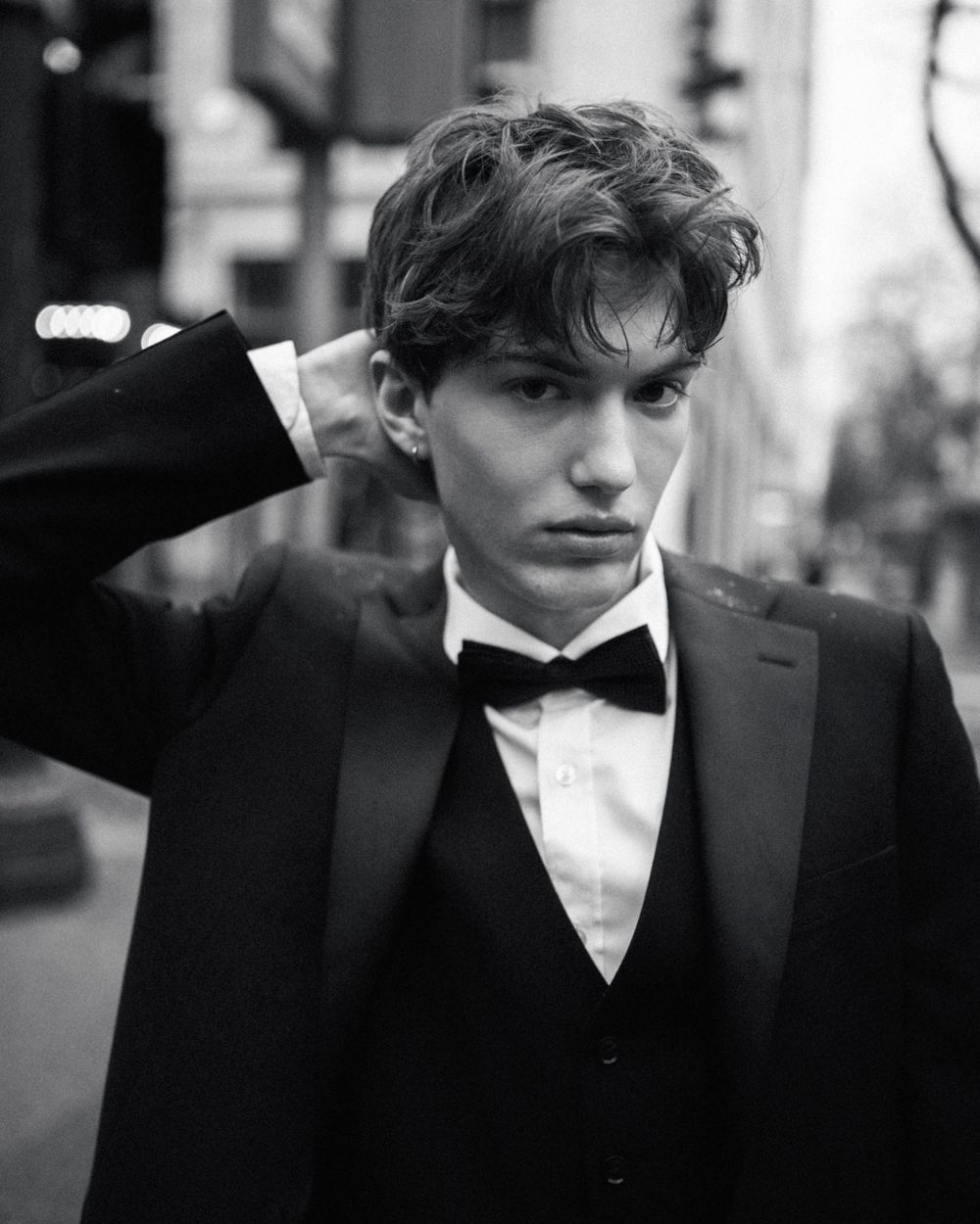 Young man in black tuxedo and bow tie poses confidently on city street in black and white photo.