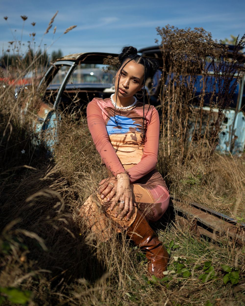 A person wearing a pink and blue outfit sits among tall dried grass near a vintage car in outdoor sunlight cinematic photoshoot.