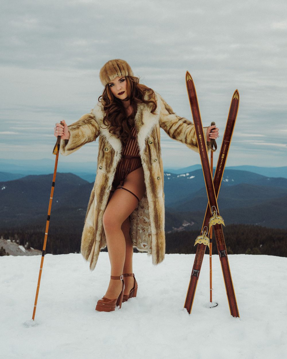 Woman in vintage fur coat and hat poses with wooden skis on snowy mountain peak.
