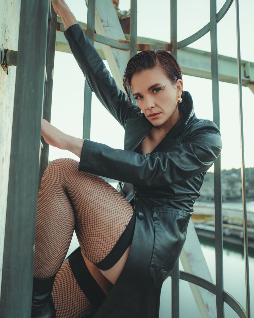 A figure in a black satin jacket and fishnet stockings poses dramatically on metal scaffolding or railings outdoors.