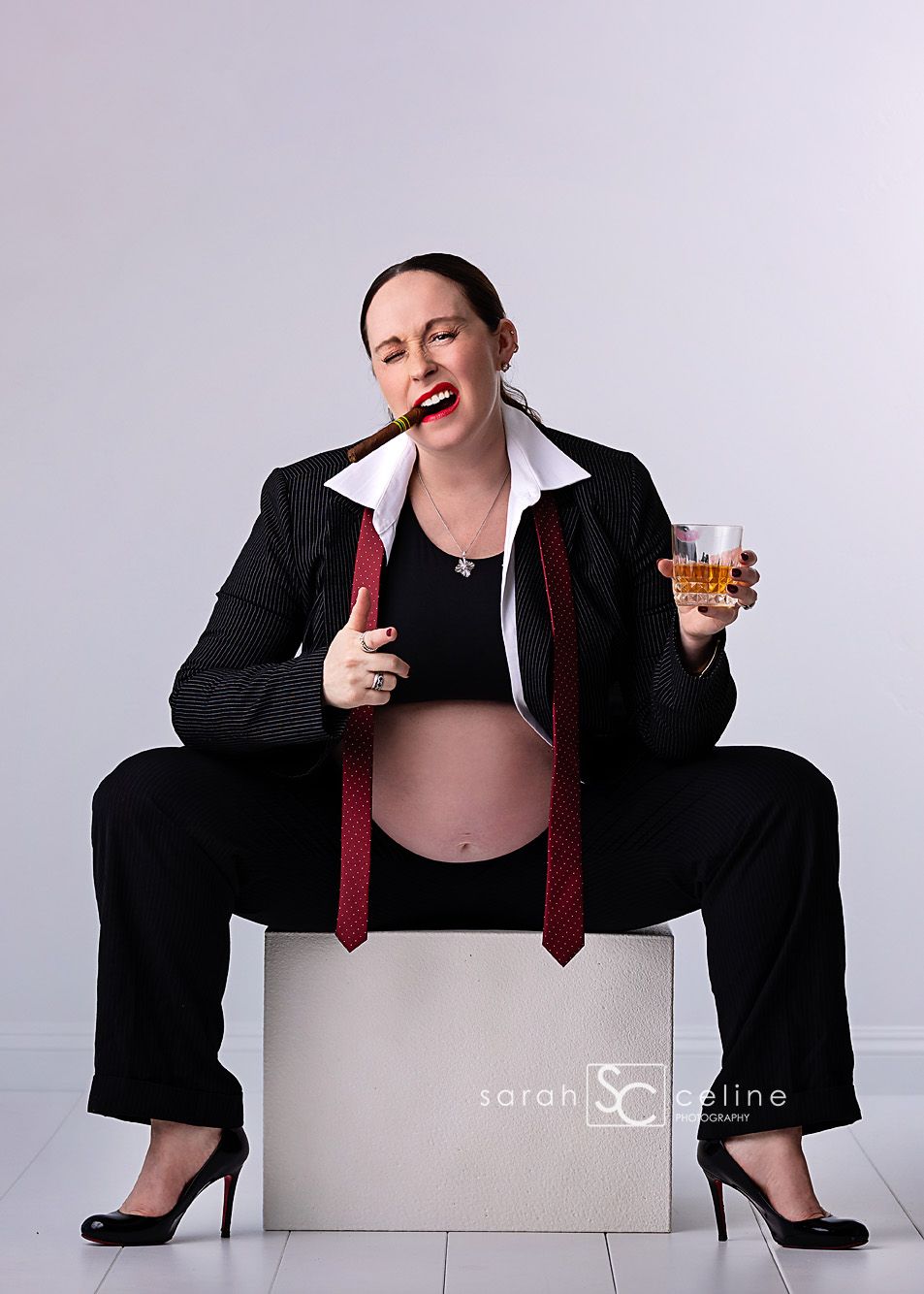 Pregnant woman in black suit holding whiskey glass and cigar, maternity photo shoot with humorous theme.