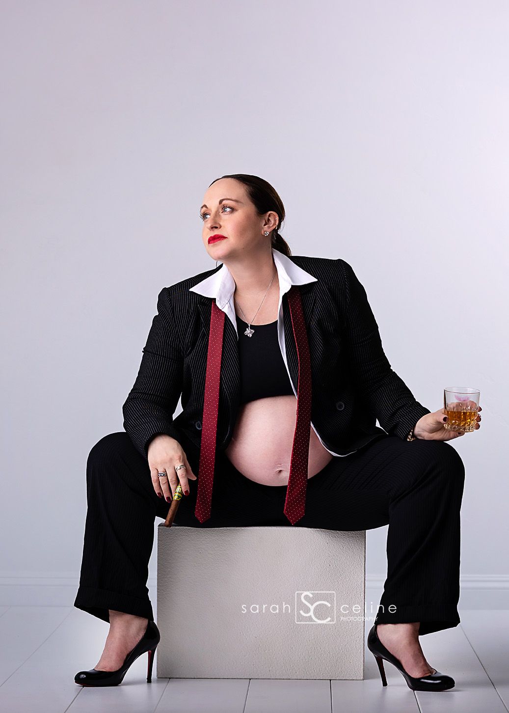 Pregnant woman in black suit with red tie holding cigar and whiskey glass, maternity photo shoot.