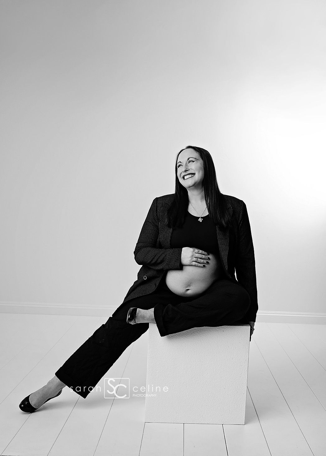 Black and white maternity photo of smiling pregnant woman in black suit sitting on white cube.
