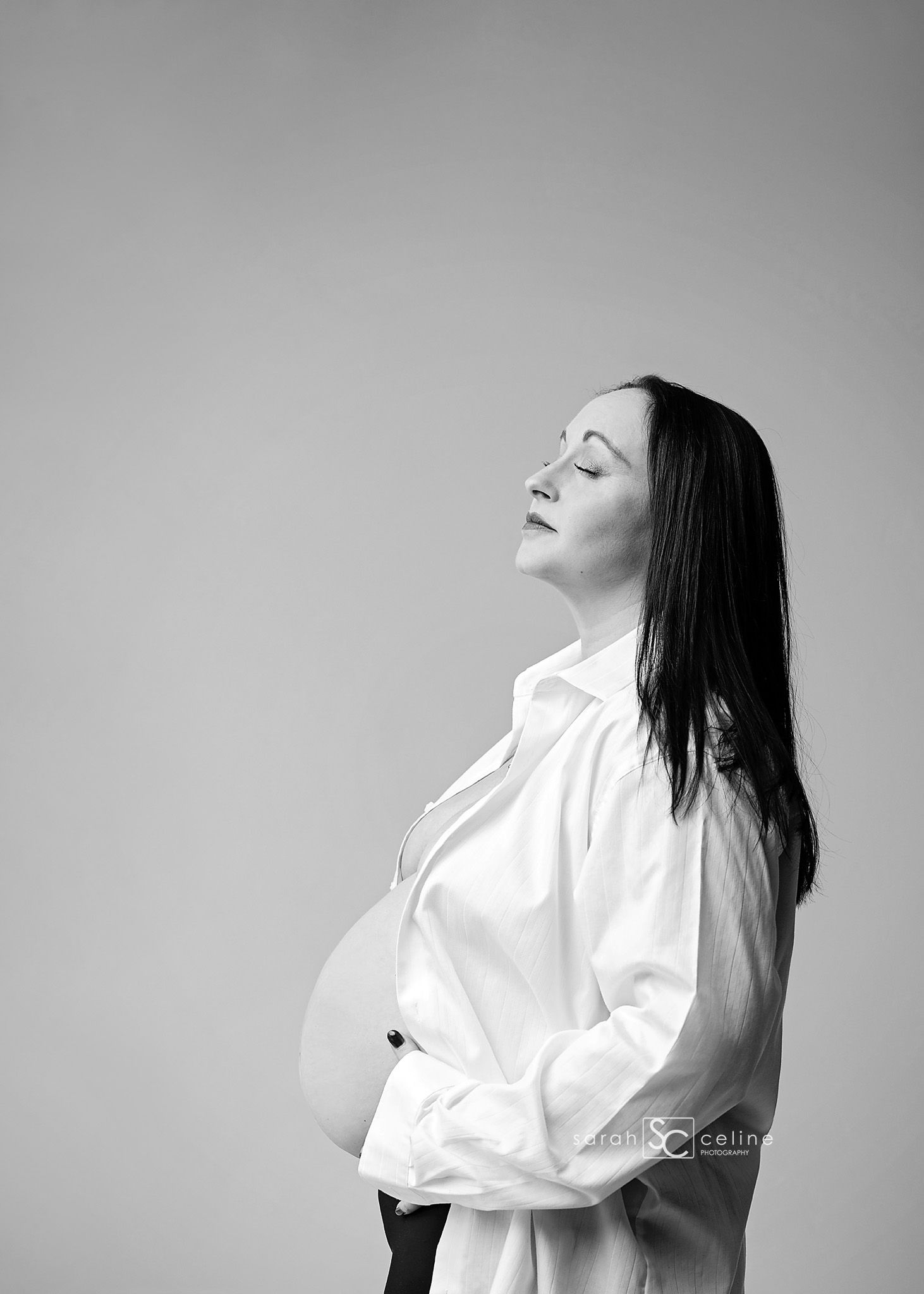 Black and white maternity photo of a pregnant woman in a white shirt, eyes closed, cradling her baby bump.