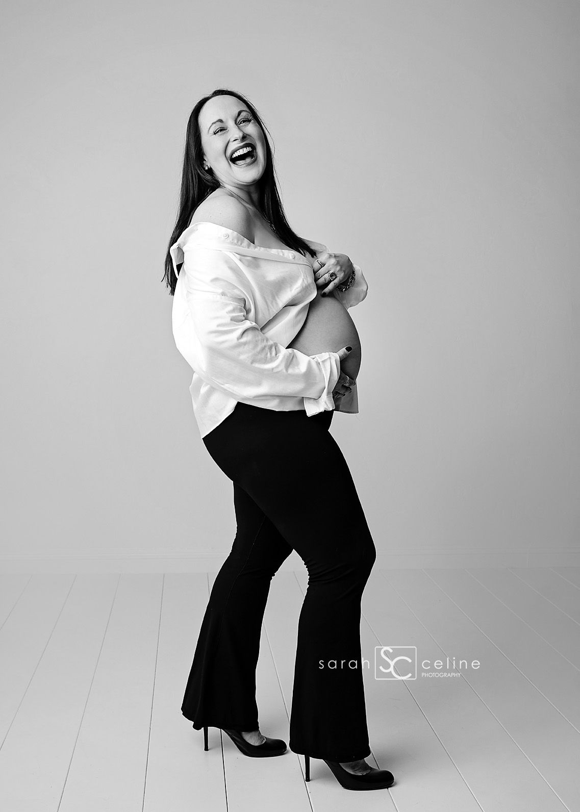 Laughing pregnant woman in white off-shoulder top and black flare pants poses sideways showing baby bump in black and white photo.
