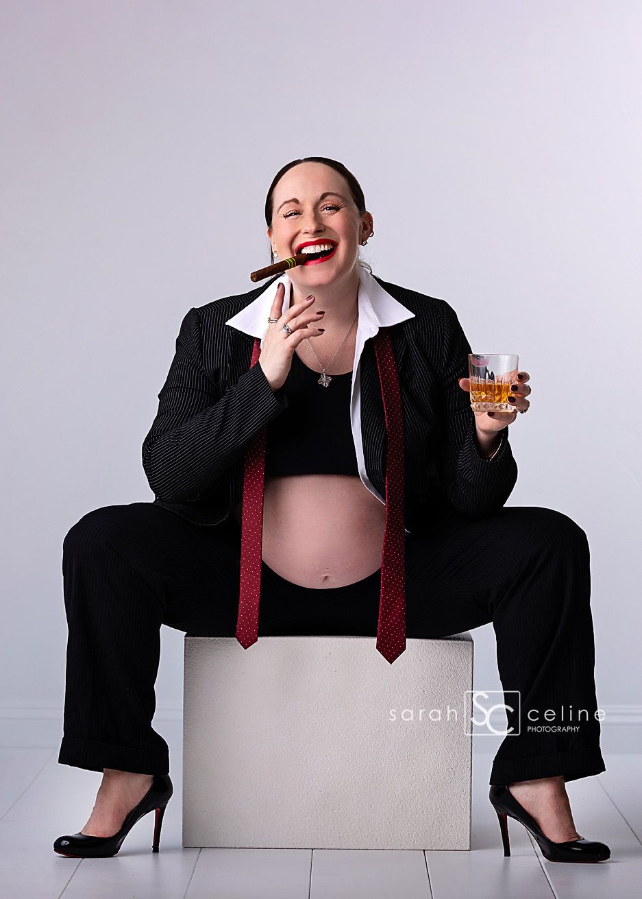 Laughing pregnant woman in black suit holding cigar and whiskey glass during fun maternity photo shoot.