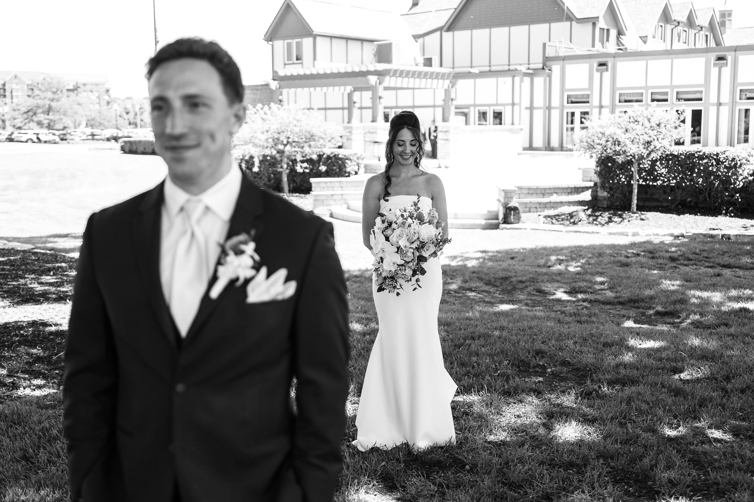 Black and white wedding photo of groom in suit and bride in white gown holding bouquet outside elegant venue.