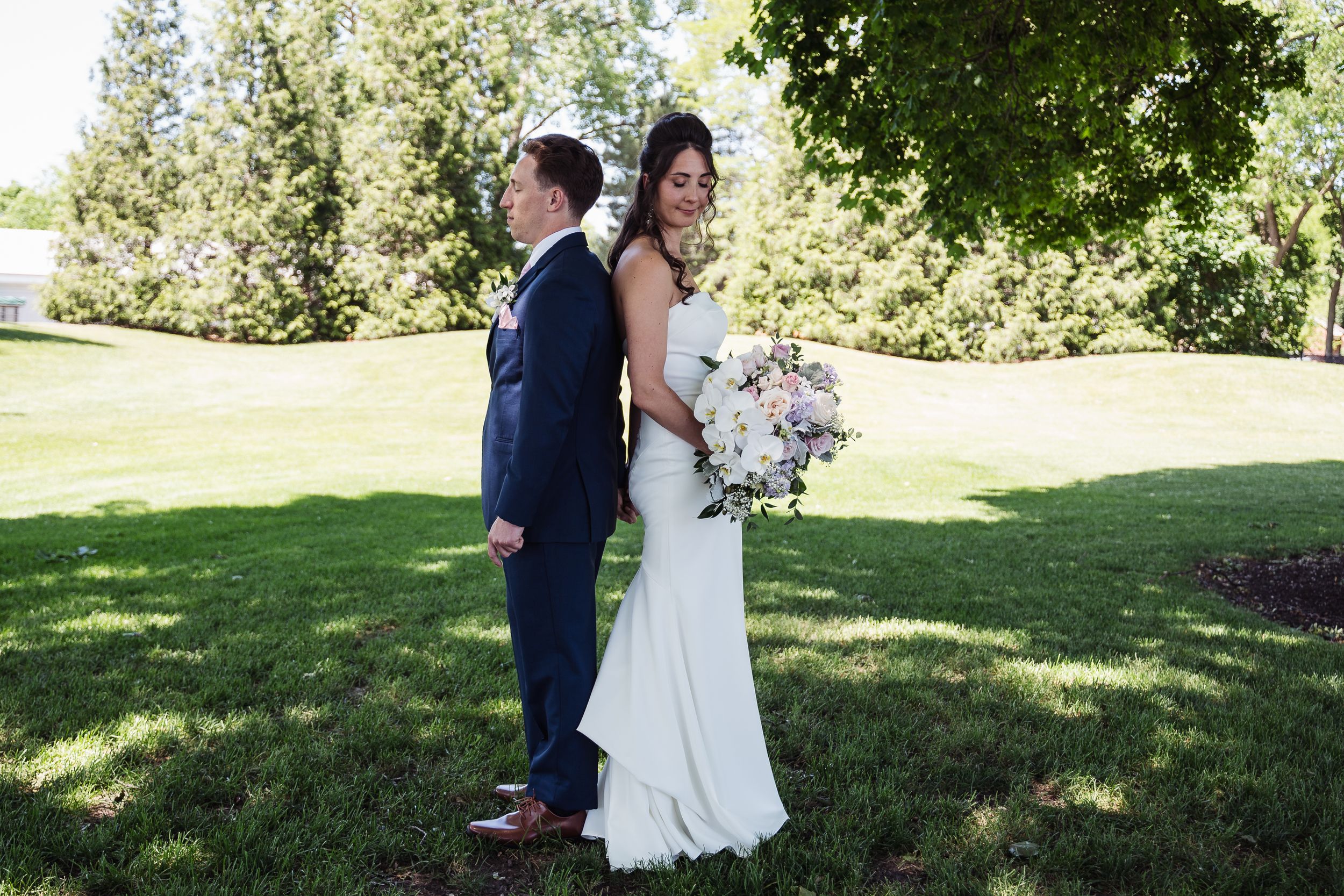 Bride in white gown holding bouquet stands back-to-back with groom in navy suit on green lawn.