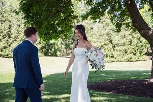 Bride in white gown holding bouquet shares first look moment with groom in navy suit outdoors.