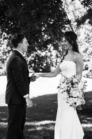 Black and white photo of a smiling bride in a strapless gown holding a bouquet while facing her groom outdoors.
