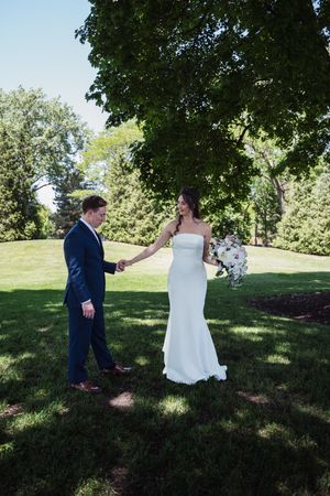 A bride in a white gown and groom in a navy suit hold hands outdoors under a large tree on their wedding day.