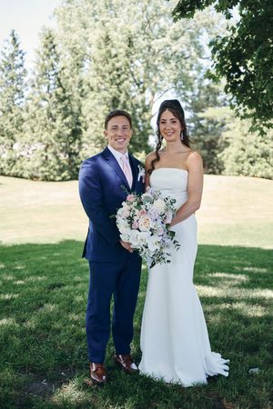 A smiling bride in a white strapless gown holds a pink bouquet beside a groom in a navy suit outdoors.