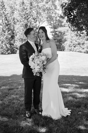 Black and white photo of a smiling bride holding a bouquet and groom embracing outdoors on their wedding day.