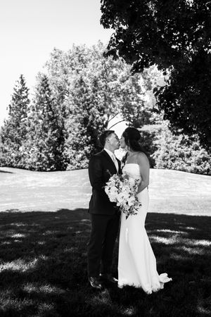 Black and white photo of a bride and groom kissing outdoors, surrounded by lush trees, bride holding a floral bouquet.