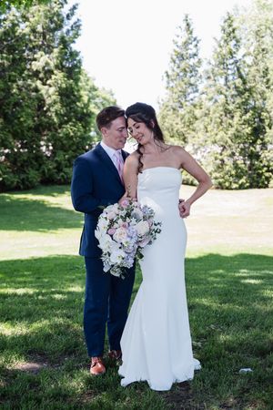 Bride in white strapless gown holding pastel bouquet stands with groom in navy suit outdoors on green lawn.