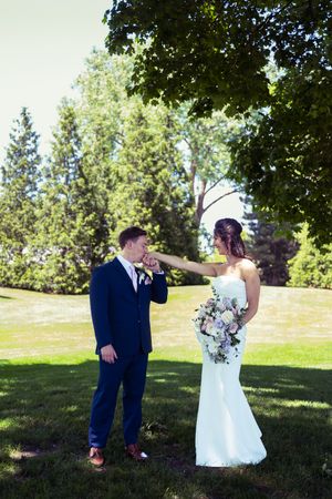 Groom in navy suit kisses bride's hand outdoors, as she holds a white floral bouquet in a lush green park.